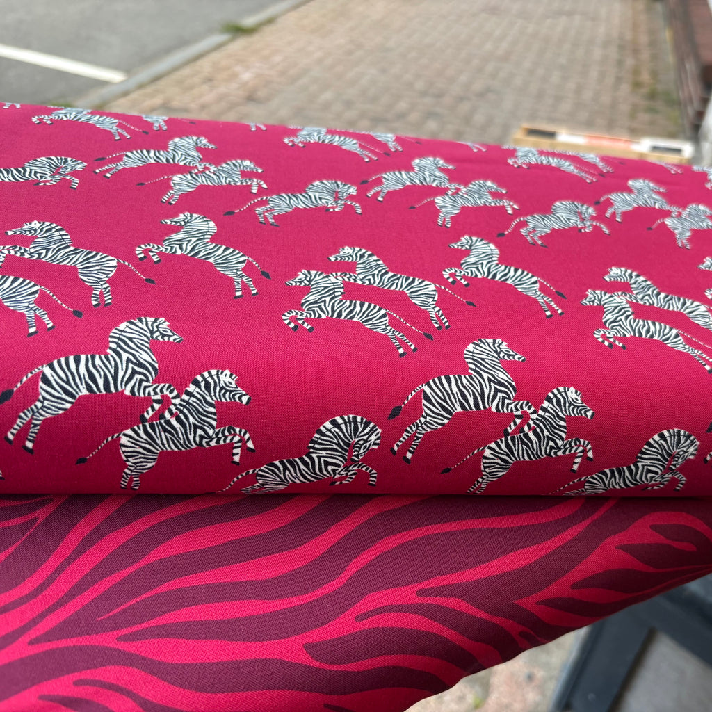 Two bolts of fabric, the one on top is magenta red background with 3 inch zebras leaping across the fabric.